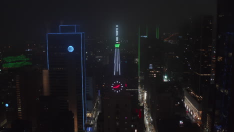 Backwards-reveal-and-tilt-up-of-neon-illuminated-spike-and-clock-on-top-of-Mercantile-National-Bank-Building.-Aerial-drone-night-view.-Dallas,-Texas,-US