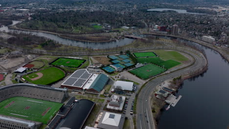 Aerial-panoramic-view-of-multilane-road-winding-on-Charles-river-waterfront-around-sports-area.-Tilt-up-reveal-of-residential-suburbs.-Boston,-USA