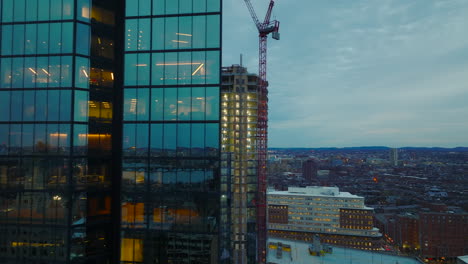 Tight-flight-around-glossy-glass-facade-of-modern-high-rise-office-downtown-building.-Revealing-illuminated-construction-site-of-new-office-tower.-Boston,-USA