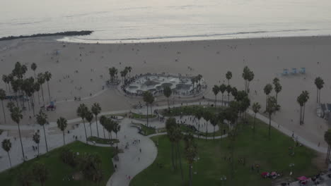 AERIAL:-Flying-away-from-Venice-Beach-Skatepark-with-Visitors-and-Palm-Trees,-Sunset-in-Los-Angeles