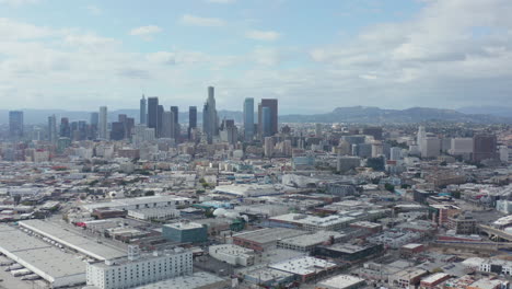 AERIAL:-Slow-Side-Shot-of-Downtown-Los-Angeles-Skyline-with-Warehouse-Art-District-in-Foreground-with-Blue-Sky-and-Clouds