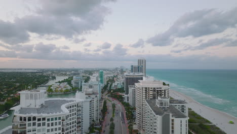 Landing-footage-of-modern-urban-borough-at-sea-coast-at-dusk.-Row-of-luxurious-multistorey-apartment-buildings-along-wide-road.-Miami,-USA
