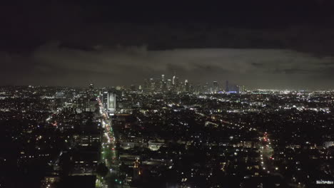 AERIAL:-Over-Dark-Hollywood-Los-Angeles-at-Night-view-on-Wilshire-Blvd-with-Clouds-over-Downtown-and-City-Lights