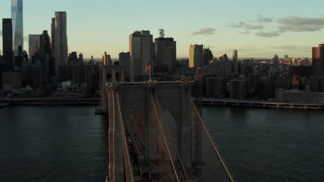 US-Flag-waving-on-top-Brooklyn-Bridge-at-sunset.-Amazing-panning-view-of-modern-high-rise-downtown-buildings.-Manhattan,-New-York-City,-USA