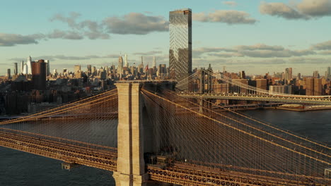 Aerial-view-of-historic-cable-stayed-bridge-over-river.-Brooklyn-Bridge-and-cityscape-in-background-lit-by-late-afternoon-sun.-Manhattan,-New-York-City,-USA