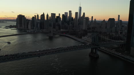 Aerial-footage-of-large-suspension-bridges-over-East-river-after-sunset.-Tilt-up-reveal-of-skyline-with-modern-tall-downtown-skyscrapers-against-colourful-sky.-Manhattan,-New-York-City,-USA