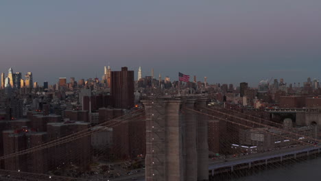 Fly-around-supporting-tower-of-Brooklyn-Bridge-with-raised-American-flag-at-dusk.-Illuminated-tall-buildings-in-distance.-Manhattan,-New-York-City,-USA