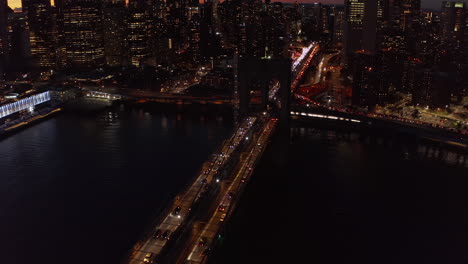 Nachts-über-Die-Brooklyn-Bridge-Fliegen.-Langsam-Fahrende-Fahrzeuge-Auf-Großer-Brücke-über-Den-Fluss.-Beleuchtete-Fenster-Von-Hochhäusern-Am-Wasser.-Manhattan,-New-York-City,-Vereinigte-Staaten