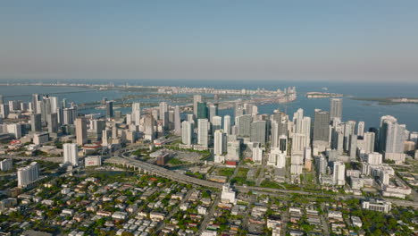 Slide-and-pan-aerial-shot-of-complex-of-tall-modern-apartment-buildings-along-coast.-Sea-bay-with-islands-in-background.-Miami,-USA