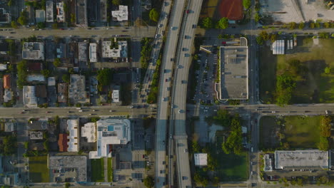 Top-down-descending-footage-of-heavy-traffic-on-multilane-highway-leading-above-urban-neighbourhood.-Golden-hour-scene.-Miami,-USA