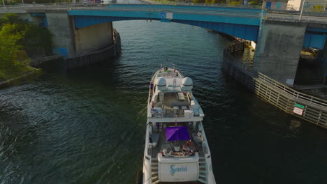 Forwards-tracking-of-luxury-yacht-passing-under-road-bridge.-People-enjoying-sunny-afternoon-on-boat-deck-floating-on-river-leading-through-city.-Miami,-USA