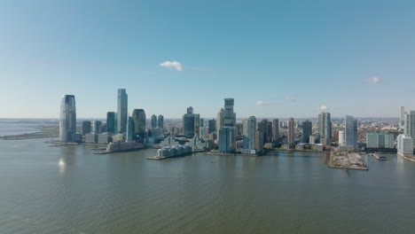 Forwards-fly-above-water.-Panoramic-shot-of-downtown-buildings-on-riverbank.-Modern-tall-skyscrapers.-Jersey-City,-USA