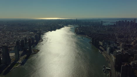 Fly-high-above-East-river-passing-between-town-districts.-Water-surface-reflecting-sunshine.-Manhattan,-New-York-City,-USA