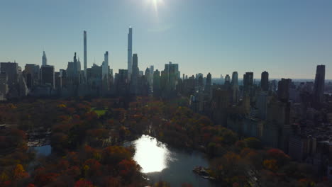 Fly-above-autumn-Central-park-with-colourful-trees-and-water-surface-reflecting-sun.-Modern-midtown-office-towers-in-background.-Manhattan,-New-York-City,-USA