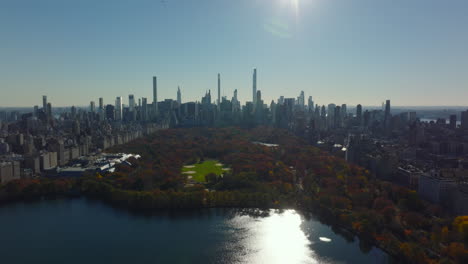 Aerial-descending-footage-of-water-reservoir,-softball-fields-and-autumn-trees-in-Central-Park-surrounded-by-high-rise-buildings.-Manhattan,-New-York-City,-USA