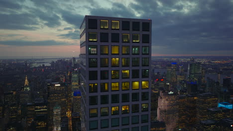 Fly-around-top-of-office-skyscraper-at-dusk.-Downtown-high-rise-buildings-with-lighted-windows-and-twilight-sky-in-background.-Manhattan,-New-York-City,-USA