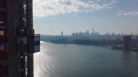 Vorwärts-Fliegen-Entlang-Der-Hochhausbaustelle.-Bauaufzug-Bewegt-Sich-Um-Die-Wand-Nach-Unten.-Wolkenkratzer-In-Der-Innenstadt-In-Der-Ferne.-Manhattan,-New-York-City,-Vereinigte-Staaten