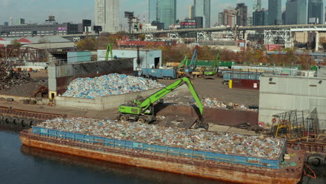 Aerial-ascending-footage-of-green-loader-standing-on-waterfront-near-barge-loaded-with-plastic-waste.-Recycling-facility.-New-York-City,-USA