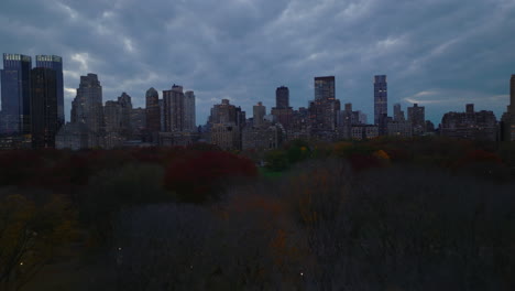 Forwards-fly-above-autumn-colour-trees-in-Central-park-at-dusk.-Panoramic-view-of-high-rise-buildings-around-urban-park.-Manhattan,-New-York-City,-USA