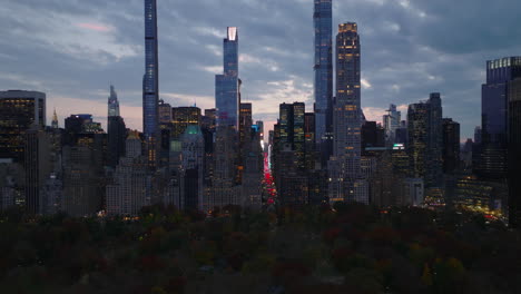Amazing-footage-of-evening-downtown.-Fly-above-trees-in-Central-park,-high-rise-buildings-and-long-straight-avenue-between.-Red-brake-lights-of-road-traffic.-Manhattan,-New-York-City,-USA