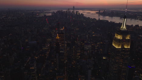 Fly-around-illuminated-upper-part-and-spire-of-Empire-State-Building.-Cityscape-at-dusk-against-colourful-sky.-Manhattan,-New-York-City,-USA