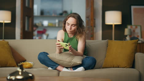 Relaxed-girl-eating-snacks-at-cozy-couch.-Carefree-woman-browsing-mobile-phone