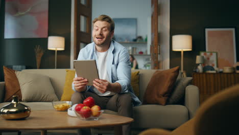 Friendly-man-waving-hand-speaking-tablet-computer-at-home.-Relaxed-guy-sitting