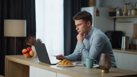 Serious-man-searching-internet-working-remotely-on-kitchen.-Young-freelancer-.