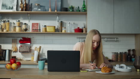 Estudiante-Que-Estudia-En-Línea-Sentada-En-La-Cocina-Con-Una-Computadora-Portátil.-Mujer-Tomando-Notas.