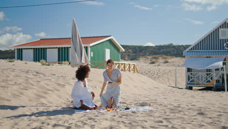 Pareja-De-Lesbianas-Usando-Un-Teléfono-Inteligente-Revisando-Fotografías-De-La-Playa.-Dos-Niñas-Descansando-En-La-Orilla