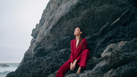 Woman-sitting-rock-ledge-in-red-suit.-Stylish-girl-looking-ocean-view-vertically