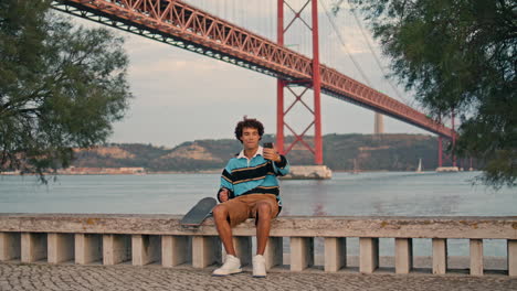 Curly-skater-making-selfie-at-coast-vertical.-Tourist-sitting-mobile-at-river