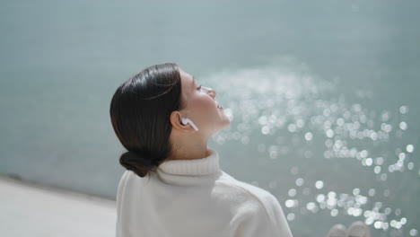 Woman-putting-face-sunlight-sitting-on-waterfront-close-up.-Girl-listening-music