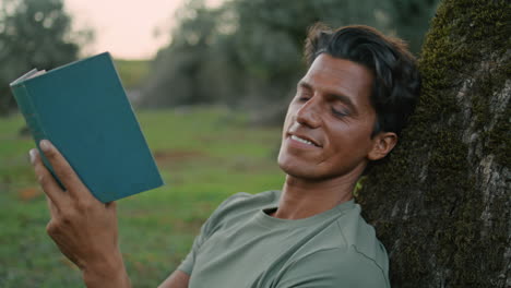 Chico-Sonriente-Sosteniendo-Un-Libro-Retrato-Del-Parque-Al-Atardecer.-Hombre-Mirando-Cámara-Leyendo-Novela