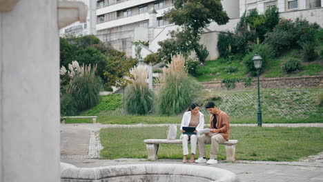 Students-couple-studying-park-bench-vertically.-Coworkers-working-tablet-talking