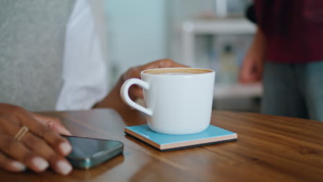 Barista-putting-cup-coffee-on-table-to-woman-sitting-with-smartphone-closeup.