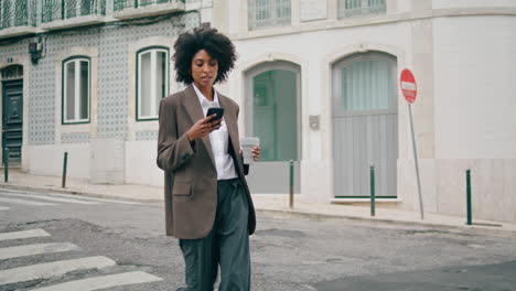 Woman-crossing-city-road-holding-cup-coffee.-african-american-lady-walking-town.