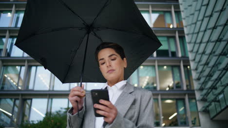Elegant-worker-texting-phone-under-umbrella.-Downtown-employee-make-online-order