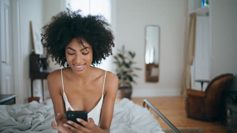 Smiling-woman-messaging-smartphone-at-bedroom.-Curly-laughing-lady-sitting-bed