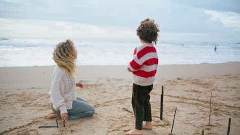 Mutter-Und-Kind-Ruhen-Sich-An-Einem-Windigen-Tag-Am-Meeresufer-Aus.-Schöne-Familie,-Die-Strandsand-Zeichnet