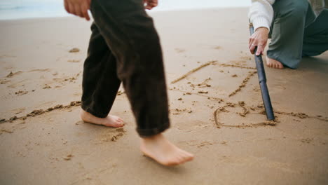 Primer-Plano-Dibujar-A-Mano-Arena-De-Playa-En-Fin-De-Semana-Familiar.-Piernas-De-Niño-Desconocido-Caminando-Junto-Al-Mar