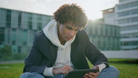 Lockiger-Freiberufler,-Der-Mit-Einem-Tablet-Auf-Dem-Rasen-In-Der-Nähe-Arbeitet.-Ruhiger-Blogger,-Der-Auf-Dem-Rasen-Sitzt