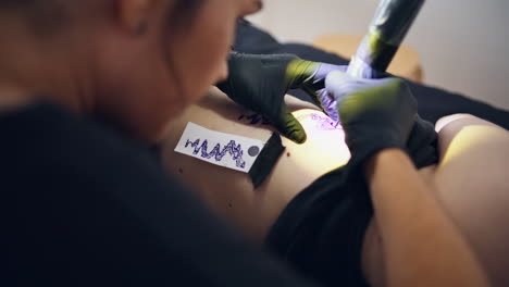 Focused-tattooer-working-machine-on-client-body-closeup.-Woman-drawing-outline