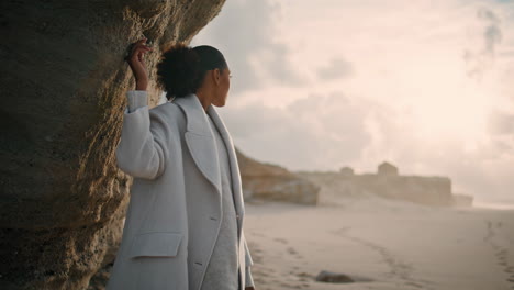 Beautiful-girl-touching-cliff-wall-on-empty-beach.-Serene-african-american-enjoy