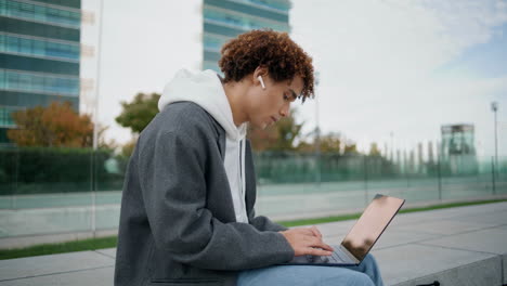 Adolescente-Rizado-Escribiendo-Una-Computadora-Portátil-En-El-Primer-Plano-De-La-Calle.-Joven-Escuchando-Música-Solo