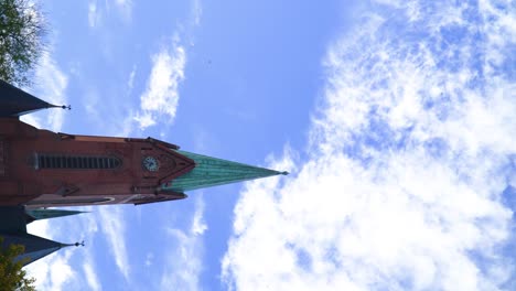 Video-Vertical-De-Una-Iglesia-Gótica-Verde-Roja-Gigante-Con-Relojes-Torres-Pequeñas-Alrededor-De-La-Parte-Superior-Puntiaguda-Del-Edificio-Histórico-En-Chequia-árboles-De-Un-Pequeño-Pueblo-Cielo-Azul-Nubes-Suaves-Que-Se-Elevan-Hacia-El-Cielo-Mágico