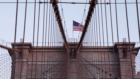 Vertical-Suspender-Cables-And-Tower-Of-Brooklyn-Bridge-In-New-York,-USA-With-American-Flag-Atop