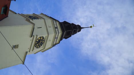 vertical-shot-of-tower-hall-with-black-and-gold-roman-numbers-historic-clocks-pointed-roof-with-golden-tiny-flag-and-small-ball-city-centre-authentic-building-soft-clouds-moving-up-Czech-republic