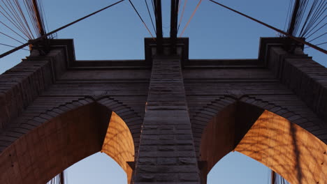 Arches-and-vertical-cables-of-Brooklyn-bridge-during-golden-sunrise,-tilt-up