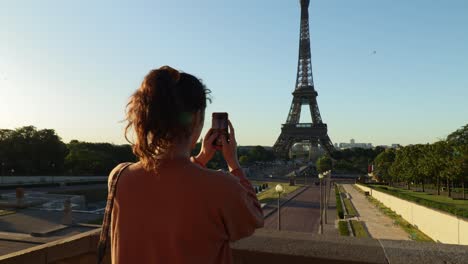 Mujer-Joven-Tomando-Fotos-Verticales-De-La-Torre-Eiffel-Con-Su-Teléfono-Durante-La-Mañana-Temprano-En-Verano,-Tiro-De-Arco-Desde-Atrás